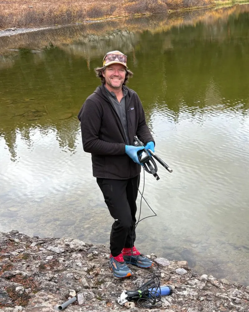 Stephen Conklin at the edge of a lake in the Lower Ice Lake Basin, holding a water-quality meter in gloved hands, with sampling gear on the rocks